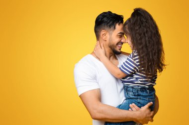 Baba Kız Bağlantısı. Küçük kızıyla bağ kuran mutlu bir Orta Doğulu babanın portresi, onun ellerini tutarak ve burnuna dokunarak sarı arka planda birlikte poz vererek,