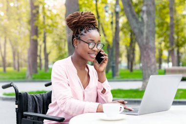 Tekerlekli sandalyedeki engelli genç bayan dizüstü bilgisayar kullanıyor, sonbahar parkındaki açık kafede akıllı telefondan sohbet ediyor. Çevrimiçi iş, engelliler için serbest çalışma