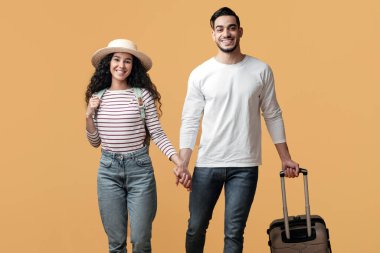 Tatil zamanı. Orta Doğulu genç bir çift, sarı arka planda yürürken, bavullu, el ele tutuşan ve kameraya gülümseyen mutlu Arap eşleri,