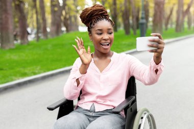 Tekerlekli sandalyedeki mutlu siyah bayan akıllı telefondan video görüşmesi yapıyor, sonbahar parkında kameraya el sallıyor. Uzaktan haberleşen neşeli özürlü Afro kadın dışarıda online toplantıya katılıyor.