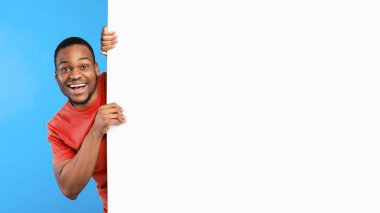 Harika bir teklif reklamı. Büyük beyaz posteri olan heyecanlı Afrikalı Amerikalı adam mavi stüdyonun arka planında kameraya gülümsüyor. Kopya Alanlı Parlak Reklam Afişi. Mockup, Panorama