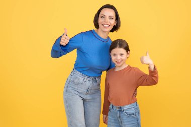 Bir anne ve reşit olmayan kızı büyük bir gülümseme sergiliyorlar ve güçlü bağlarını ve sevgilerini gösteriyorlar. Enerji ve neşe dolu, canlı sarı bir arka plana karşı dururlar..