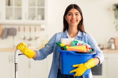 Ev işleri. Elinde kovayla temizlik malzemeleri ve paspas taşıyan, gülümseyen ve mutfakta duran kameraya bakan neşeli bir ev kadını portresi. Profesyonel temizlik servisi uzmanı yardım öneriyor