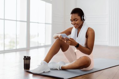 Omzunda havlu ve kablosuz kulaklığı olan mutlu Afrikalı bir kadın oturma odasında oturmuş, dinlenirken müzik dinliyor, bedava fotokopi alanı. Spor müziği mobil uygulama kavramı