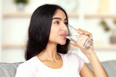 Susuz kalma konseptini koru. Çekici genç Hintli kadın oturma odasındaki kanepede camdan su içiyor. Güzel Hindu bayan sağlığına ve sağlığına dikkat ediyor.