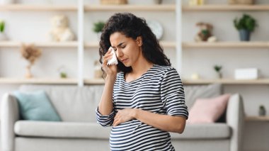 Hamilelik hormonları konsepti değiştirir. Genç kıvırcık saçlı hamile kadın kendini kötü hissediyor, rahat bir oturma odasında yerde oturuyor ve ağlıyor, peçete kullanıyor, evde tek başına kalıyor, boş bir alanda.