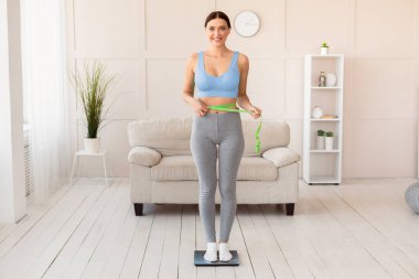 Başarılı Kilo Verme. Gülümseyen kız, ince belini ölçüyor, kilolarını evde ölçüyor, kameraya bakıyor. Tam Uzunluk, Boş Alan