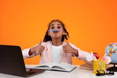 Burun ve dudak arasında kalem tutan komik zenci kız öğrencinin portresi, baş parmak işareti hareketi gösteren, sarı stüdyo arka planında bilgisayarla masada oturan, bedava fotokopi alanı