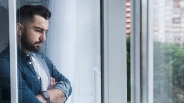Depresyondaki beyaz adam işini kaybediyor ve kalbi kırık, pencerenin yanında yalnız. Travma sonrası stres bozukluğu, akıl sağlığı ve depresyon. Mutfaktaki ıslak pencereye bakan kollarını kavuşturmuş üzgün, yakışıklı genç adam.