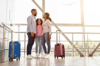 Family Travel Concept. Portrait of black dad, mum and girl standing at departure lounge with suitcases waiting for the aircraft arrival, looking at each other and hugging. Free copy space