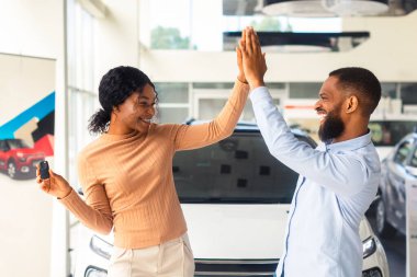 Neşeli Afrikalı Amerikalı çift, yeni araba aldıktan sonra birbirlerine beşlik çakıyor, gösteriş salonunda duran ve anahtarı tutan heyecanlı siyah eşler, aile aracını, bedava alanı kutluyorlar.