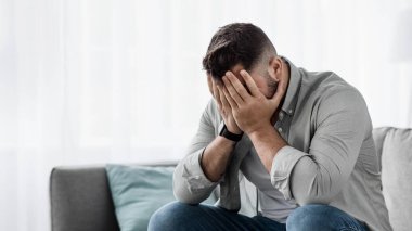 Utanç verici umutsuzluk, üzgün adamın portresi baş ağrısı, stres ve depresyondan muzdarip. Bin yıllık mutsuz Avrupalı yakışıklı adam ağlıyor, yüzünü ellerle kapatıyor, oturma odasındaki kanepede oturuyor.