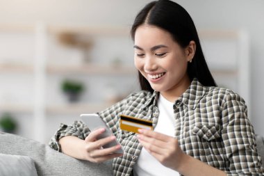 Modern akıllı telefon ve kredi kartı tutan neşeli genç Çinli kadın, evde otururken internet üzerinden mal ve hizmet için ödeme yapıyor, internet üzerinden alışveriş yapmak için mobil uygulama kullanıyor,
