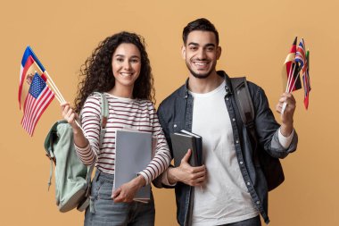 Yurtdışında eğitim. Uluslararası bayraklar taşıyan mutlu genç Arap ve kadın sırt çantaları ve çalışma kitaplarıyla gülümseyen Orta Doğulu öğrenciler sarı stüdyo geçmişine poz veriyorlar.