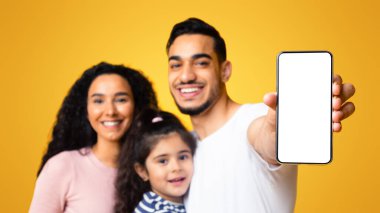 Üç kişilik mutlu bir orta doğu ailesi beyaz ekranlı boş akıllı telefonu gösteriyor. Heyecanlı bir Arap ebeveyn ve küçük kızı sarı arka planda durup yeni mobil uygulama öneriyorlar.
