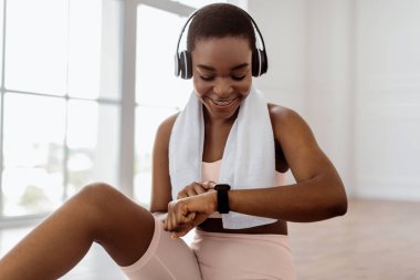 Gülümseyen Afrikalı genç bir kadın spor molasında aktivite takip cihazına bakıyor, kalp atışlarını kontrol ediyor ve kalori yakıyor. Kablosuz kulaklık, spor sutyen ve havlu takan mutlu kadın.