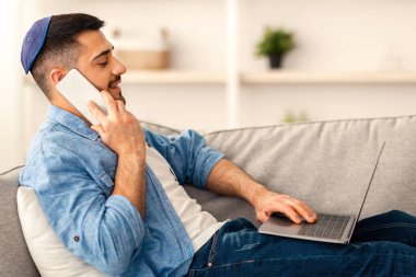 İçişleri Bakanlığı Konsepti. Bin yıllık gülümseyen bir adamın iş yerindeki bilgisayarını kullanarak koltukta otururken akıllı telefondan konuşmasının yan görünüm portresi. Genç erkek çalışan müşteriye danışıyor, arkadaşlarıyla sohbet ediyor.