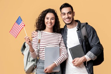 Yurtdışında oku. Gülümseyen Orta Doğulu öğrenciler, ellerinde Amerikan bayrağı, sırt çantaları ve çalışma kitapları olan mutlu Arap erkek ve kadın, sarı stüdyonun arka planında duruyorlar.