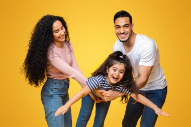 Aile eğlencesi. Neşeli Arap ebeveynlerin küçük kızlarıyla oynamaları, uçan, ellerini ayıran, anne ve babasıyla sarı arka planda oynaşan komik kadın çocuk portresi.