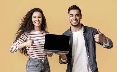 Gülümseyen Arap bir çift ellerinde siyah ekranlı dizüstü bilgisayarlarıyla başparmaklarını kaldırarak mutlu Orta Doğulu erkek ve kadınların yeni bir web sitesi önermelerini sarı arka plan üzerinde izole edilmiş bir şekilde duruyorlar.