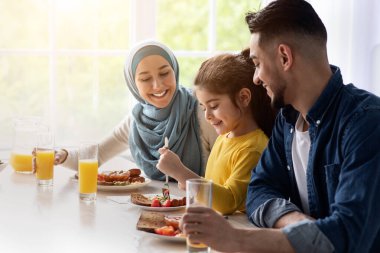 Birlikte öğle yemeği. Mutlu İslami ebeveynler ve küçük kız mutfakta lezzetli yemekler yiyor, üç kişilik neşeli, genç bir Arap aile masada oturuyor, lezzetli yemekler yiyor ve sohbet ediyor,