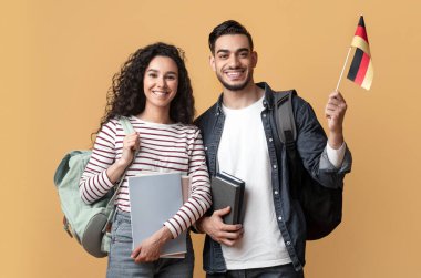 Uluslararası Eğitim. Sırt çantalı Ortadoğulu çift ellerinde Alman bayrağı, gülümseyen Arap ve kadın portresi kolej kitaplarında sarı stüdyo arka planında poz veriyorlar.