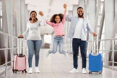 Ailesi havaalanında. Üç Afro-Amerikan 'ın eğlenceli portresi, baba ve anne atlayan kızla el ele tutuşuyor, havaalanı terminalinde birlikte bir yolculuğa çıkıyorlar.