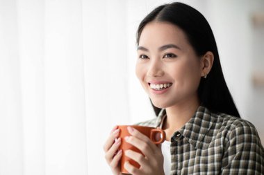 Pencereden bakan, gülümseyen, elinde kahve fincanı, elinde fotokopi odası olan Asyalı kadının yakın çekimi. Güzel Çinli kadın sabah çayının keyfini çıkarıyor, pencerenin önünde duruyor.