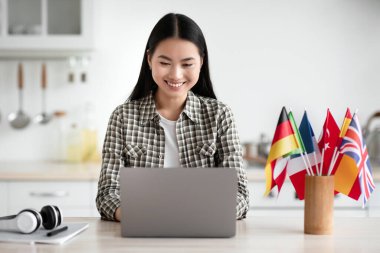 Çevrimiçi yabancı dil kursu arayan neşeli Asyalı genç kız öğrenci, mutfakta oturuyor, modern dizüstü bilgisayar kullanıyor, masada uluslararası bayraklar, online okul konsepti