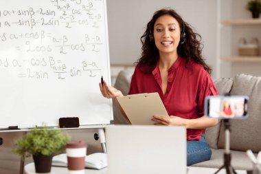 Uzaklık Eğitim Konsepti. Video çağrısı yapan öğrencilere trigonometrik fonksiyonları açıklayan matematik öğretmeninin portresi. Kulaklık takmış bir kadın dizüstü bilgisayara bakıyor, cep telefonundan kayıt dersi alıyor.
