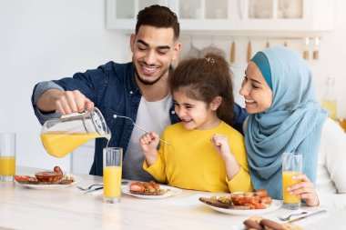 Sağlıklı içecek. Mutlu Arap baba, kızı ve karısı için portakal suyu doldururken, onlar mutfakta kahvaltı ederken, neşeli Müslüman aile evde gülümsüyor ve eğleniyorlardı.