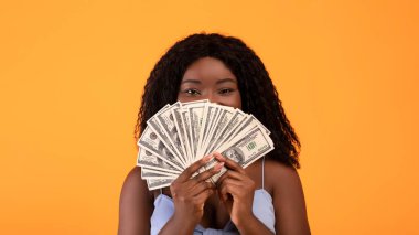 Yüzüne para fanı süren genç siyahi bayan, turuncu stüdyo arka planında alışveriş bütçesi gösteriyor. Milenyum Afrikalı Amerikalı kadın satın almak için para alıyor.