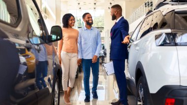 Araba satıcısı, siyahi eşlere galeride yeni otomobil seçmeleri için yardım ediyor. Profesyonel satıcı, genç Afrikalı Amerikalı aileye araç özelliklerini açıklıyor.