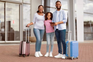 Modern havaalanı terminalinin yanında duran üç kişilik neşeli Afro-Amerikan ailesinin portresi, babamın elinde bavulları, pasaportları ve biletleri, kameralara ve poz verdirme
