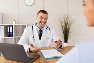 Test sonuçlarını açıklıyorum. Gülümseyen orta yaşlı bir doktorun portresi. Ofisindeki randevusu sırasında bayan hastaya danışıyor, dizüstü bilgisayarla oturuyor. Klinikteki genç bayanla konuşan bir uzman.