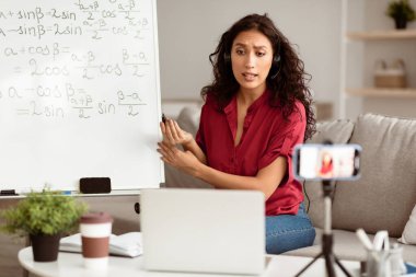 E-öğrenme. Kendine güvenen bir kadın bilgisayarla video konferans görüşmesi yapıyor. Kablosuz kulaklık takan bayan öğretmen web kamerasıyla konuşuyor, cep telefonuyla tripodla özel ders kaydı yapıyor, canlı yayın yapıyor.