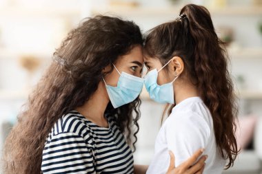 Genç esmer kadın ve koruyucu maske takan kıvırcık kız, ev içi. Anne ve çocuk koronavirüs salgını sırasında dışarı çıkıyorlar, ebeveyn çocuğunu korurken...