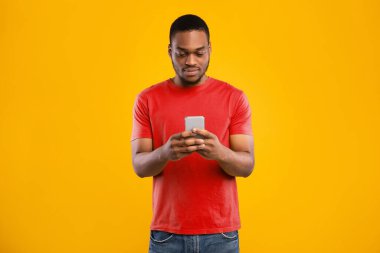Afrikalı Amerikalı Milenyum Adamı Akıllı Telefon Tarama ve Sarı Arkaplan Üzerinde Mesaj Kullanıyor. Stüdyo fotoğrafı, adamın yeni cep telefonunda uygulamayı kullanırken çekildi. Cep telefonunuz için Uygulama
