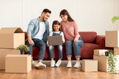 Ev Sahipliği ve Yerleştirme Projesi Konsepti. Yeni eve taşındıktan sonra dizüstü bilgisayar kullanan mutlu bir ailenin portresi. Anne, baba ve çocuk internette geziniyor, ekrana bakıyor.