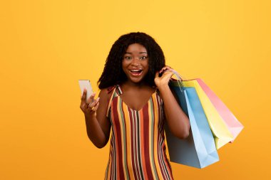 Huge online sale concept. Portrait of excited African American woman with shopping bags using smartphone, purchasing goods on web via new mobile app over orange studio background