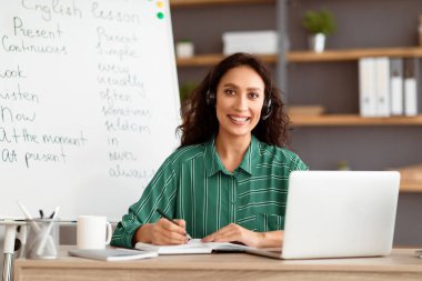 Uzaktan Çalışma Konsepti. Kablosuz kulaklıklı gülümseyen bir kadının portresi masa başında dizüstü bilgisayarla beyaz tahtanın yanında oturuyor. Mutlu bayan poz veriyor ve kameraya bakıyor, yazıyor, not alıyor.