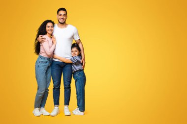 Aile portresi. Mutlu Orta Doğulu ebeveynler ve küçük kızları sarı arka planda kucaklaşırken mutlu bir Arap anne, baba ve çocuğun stüdyoda kucaklaşması.
