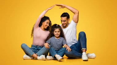 Aile koruması. Küçük kızlarının üstünde çatı yapan Orta Doğulu ebeveynler, sarı arka planda yan yana otururken Arap anne ve babalarının kadın çocukla yakınlaşması.