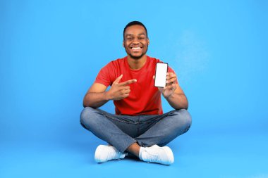 Gülümseyen Afrikalı adam cep telefonunu boş ekranla gösteriyor. Mavi Stüdyo Arkaplan 'da oturan akıllı telefon için harika bir uygulama öneriyor. People and Gadgets, Yeni Başvuru. Model
