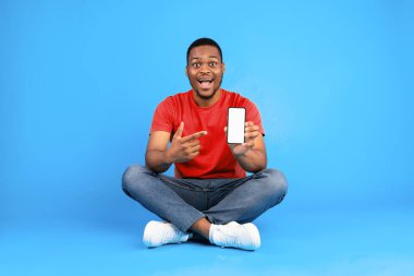Excited Black Guy Showing Smartphone Pointing Finger At Blank Cellphone Screen Advertising And Recommending New Mobile Application Sitting Over Blue Studio Background. Mockup