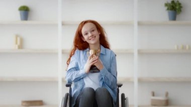 Engelli neşeli bir kızıl kız tekerlekli sandalyesinde oturmuş elinde kupa tutarken mutlu bir şekilde gülümsüyor. Dekoru az olan, iyi ışıklandırılmış bir oda, rahat bir atmosfer izlenimi veriyor..