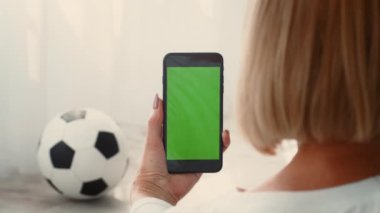 Son sınıf öğrencisi bir kadın evde spor yapıyor, akıllı telefonunu kontrol ediyor. Yakınlarda, bir futbol topu yerde duruyor, fitness ayarlarına sportif bir dokunuş ekliyor..