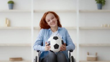 Tekerlekli sandalyedeki neşeli bir Avrupalı kız elinde bir futbol topu tutarken gülümsüyor. Parlak oda, onun pozitif ruhunu ve spora olan sevgisini yansıtan sıcak bir atmosfer sergiliyor..