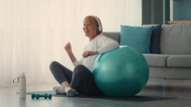 Neşeli yaşlı bir bayan evde egzersiz yapmak için stabil bir top kullanıyor. Kulaklık takıyor ve parlak, rahat bir yerde spor yapmayı seviyor..