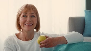 Neşeli yaşlı bir kadın evde bir fitness seansına katılıyor. Yanında dengeli bir topla egzersiz yapmaya hazırlanırken elinde sarı bir elma tutuyor..